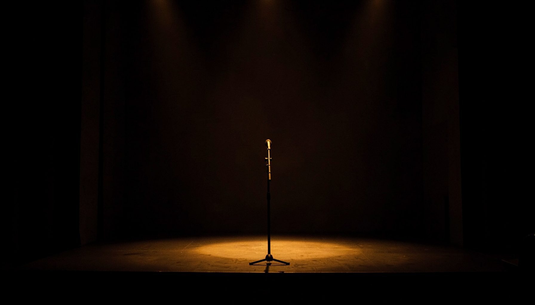 Intimate live performance setting with subtle stage lighting, warm amber glow on dark surroundings, a single microphone stand visible