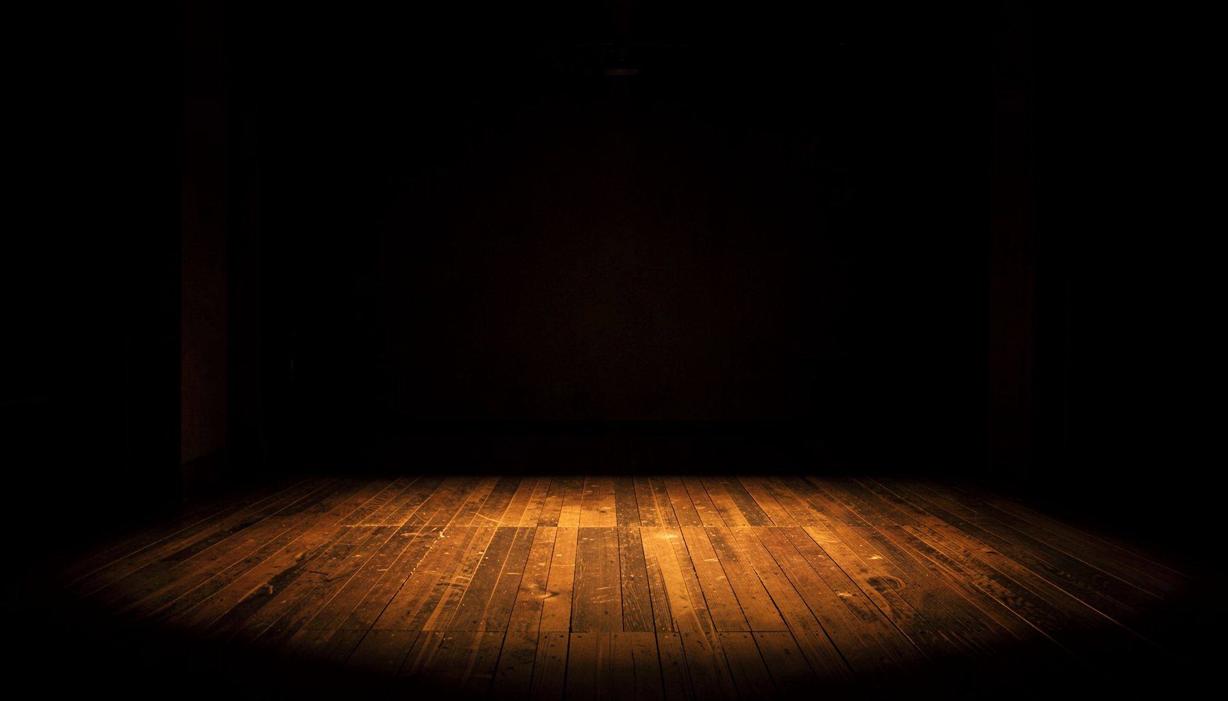 Dark atmospheric stage with warm amber light spilling across textured floorboards and deep shadows pooling at the edges, a moody and contemplative mood