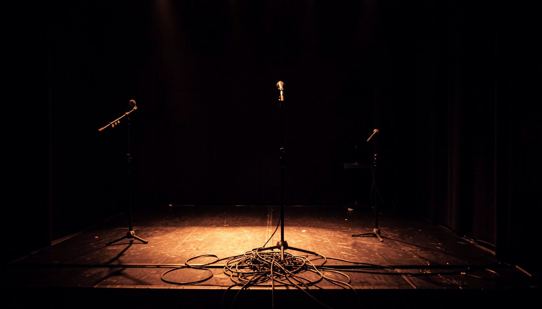 A dimly lit stage with microphone stands and tangled cables, warm amber and deep indigo shadows, an intimate performance space awaiting sound.