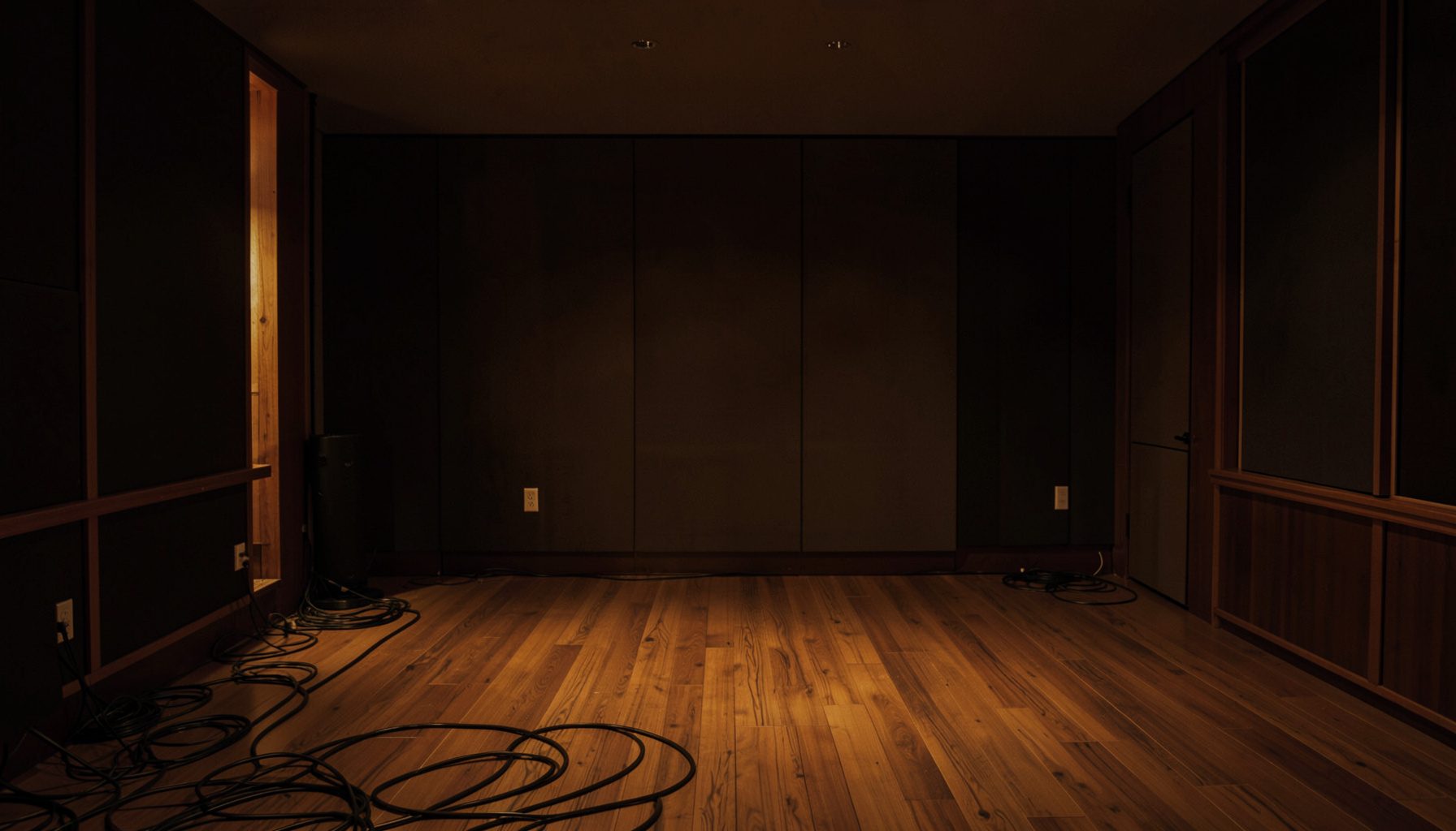 A dimly lit recording studio interior with warm amber and deep brown tones, cables trailing across a wooden floor