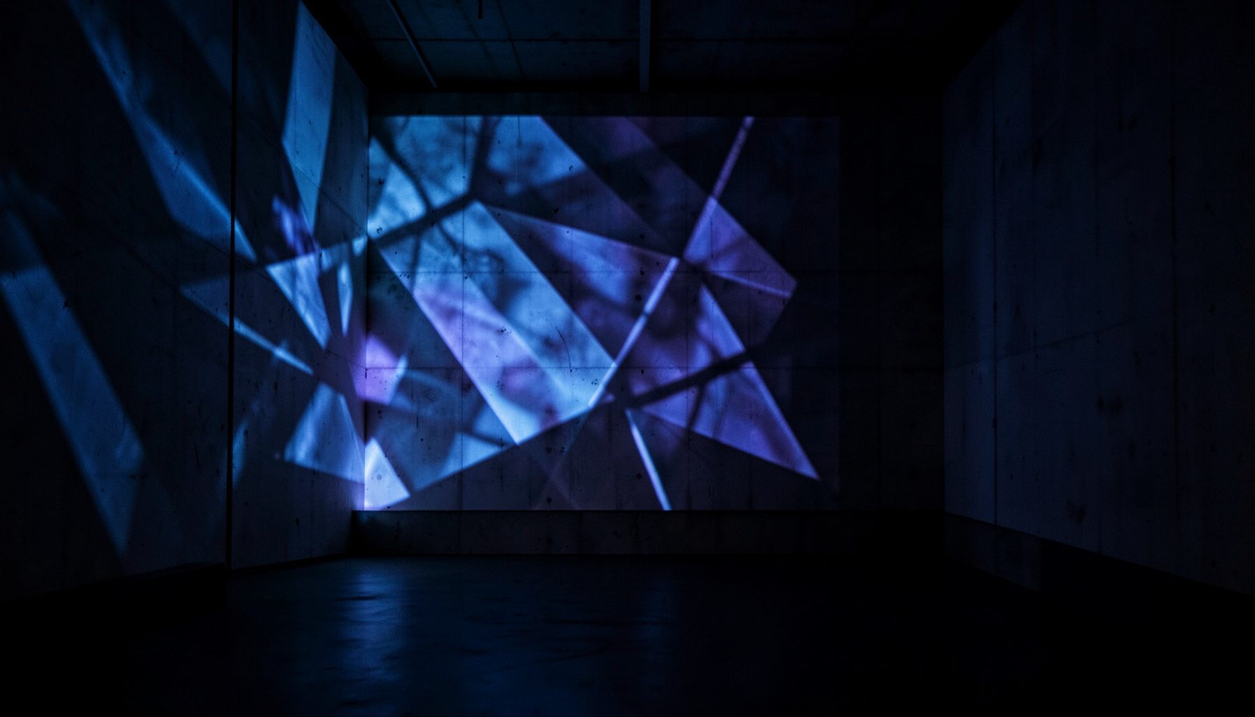 A dimly lit gallery interior with a large-scale projection casting geometric patterns across a concrete wall, cool blue and warm amber intersecting