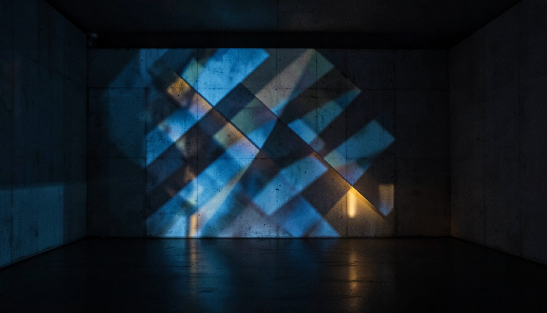 A dimly lit gallery interior with a large-scale projection casting geometric patterns across a concrete wall, cool blue and warm amber intersecting in a contemplative atmosphere